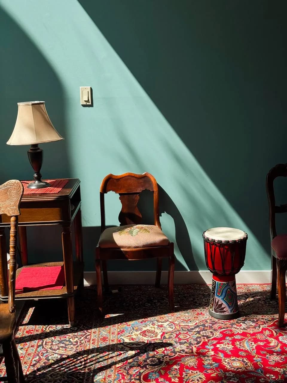 A sunlit corner: teal wall, wooden chair, lamp and a hand drum on a Persian rug