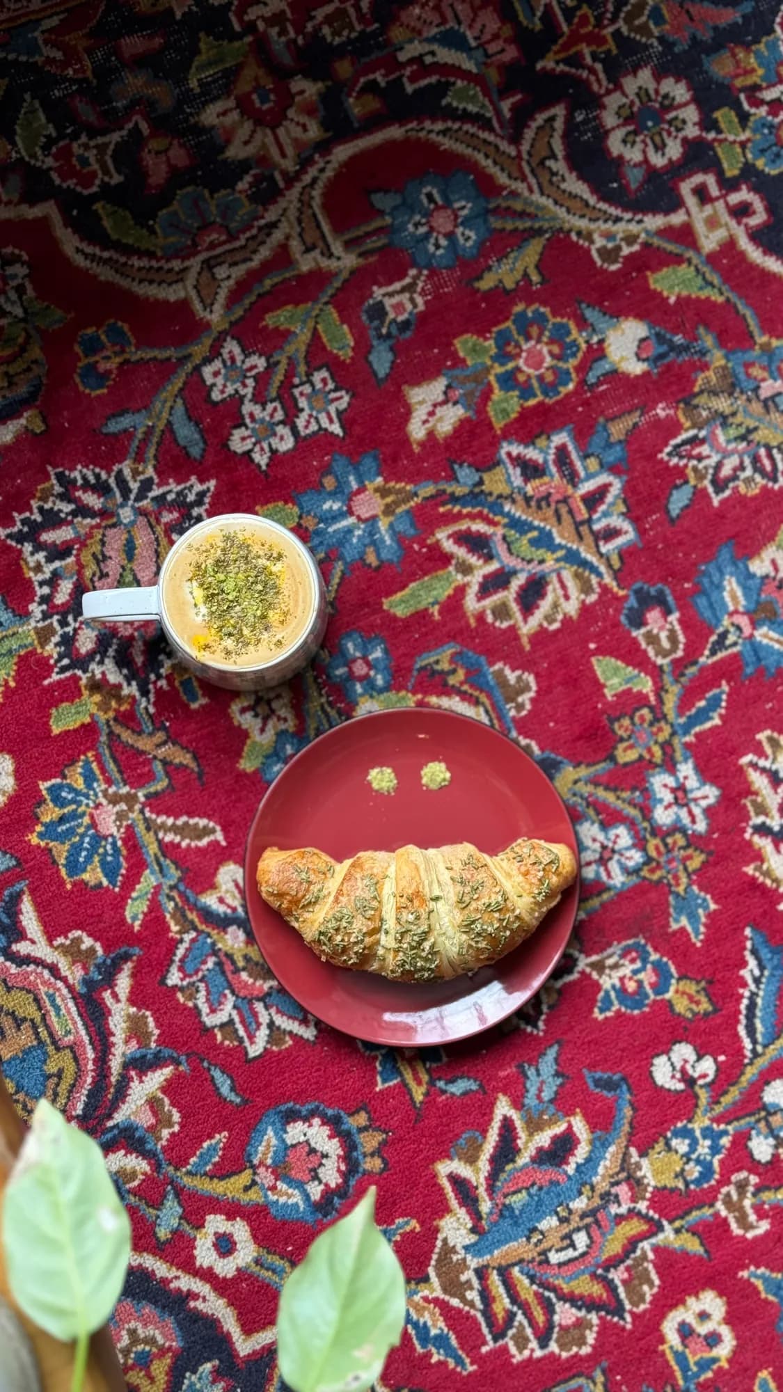 A croissant on a red plate and a matcha latte, seen from above on a Persian rug